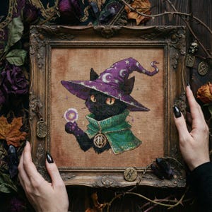 May include: Cross-stitch art of a black cat wearing a purple wizard hat with crescent moons and stars. The cat has orange eyes and a green cape, holding a crystal ball. The artwork is framed in a decorative, ornate wooden frame.