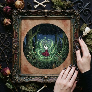 May include: A framed cross-stitch depicting a character in a red dress and white mask in a forest scene. The artwork is framed in a dark, ornate frame with a green and brown color scheme. The background includes dried roses and bones.