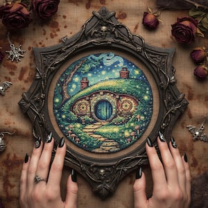 May include: A cross-stitch of a hobbit hole scene, set in a dark, ornate frame. The artwork depicts a blue door, green hills, and a starry night sky. The frame is held by hands with black nail polish.