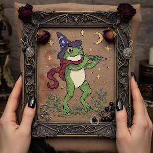 Puede incluir: Arte de punto de cruz de una rana verde tocando la flauta, con un sombrero de mago y una bufanda. La obra está enmarcada en un marco oscuro y ornamentado con elementos decorativos. Rosas secas y abalorios adornan el marco.