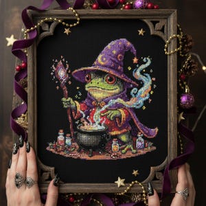 Puede incluir: Imagen de punto de cruz de un sapo mago con sombrero y túnica morados, removiendo un caldero. El sapo es verde con ojos rojos, sosteniendo un bastón. La obra está enmarcada en un marco de madera.