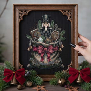 May include: Cross-stitch picture of a bag filled with festive items, including a nutcracker, pine cones, and a bottle. The bag is tied with a large red bow and framed in a dark wooden frame. Gold bells and greenery decorate the frame.