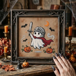 May include: A cross-stitch embroidery in a dark wooden frame featuring a white ghost wearing a silver knight helmet and a red cape, holding a gray sword. The design includes two orange jack-o'-lanterns, black bats, and a crescent moon on a tan fabric background.