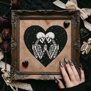 Puede incluir: Obra de arte de punto de cruz en un marco de madera decorativo. El diseño presenta dos esqueletos abrazándose dentro de un corazón negro sobre un fondo estrellado. El marco es ornamentado, con un acabado marrón oscuro. Rosas secas y elementos decorativos rodean la obra enmarcada.