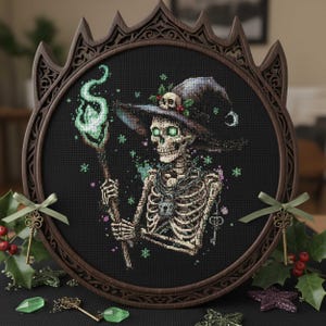May include: Cross-stitch art of a skeleton witch with a staff emitting green flames, wearing a witch hat adorned with a skull and holly. The artwork is framed in a dark brown, ornate, round frame, with decorative keys and gems.