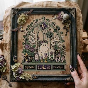 Könnte beinhalten: Eine gerahmte Kreuzstichstickerei mit einer zentralen Geisterfigur in einem Gewächshaus mit violetten und grünen Blumenmotiven, Pilzen und Zaubertrankflaschen. Der dunkle Rahmen ist mit kleinen Metallfledermäusen, Schlüsseln und einem Totenkopf verziert.