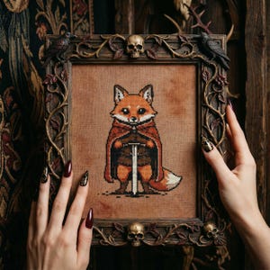 May include: Cross-stitch art of a fox wearing a red cape and holding a sword, framed in a dark wood frame with decorative skulls and foliage. The fox has orange and white fur. The frame is held by hands with long, dark red nails.