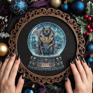 May include: A cross-stitch design featuring a black cat in a snow globe, adorned with gold and seated on a throne. The globe is set against a black background, surrounded by Christmas ornaments and a wooden frame. The text "THE CURSE OF ANUBIS" is visible.