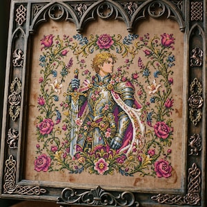 May include: Cross-stitch artwork of a knight in ornate armor, holding a sword, surrounded by pink roses and floral elements. The artwork is framed in a dark, decorative wooden frame with gothic-style accents. The knight has blonde hair and is wearing a cape.