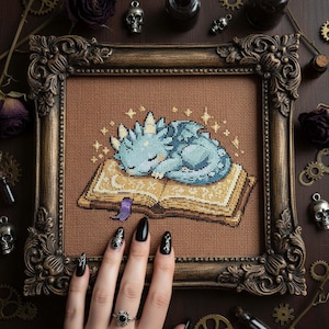 May include: Cross-stitch art of a sleeping blue dragon on an open book, framed in a decorative brown frame. The dragon is surrounded by sparkling stars. The frame is on a dark brown surface with decorative elements.