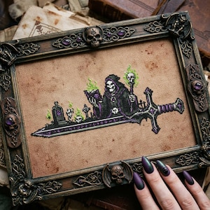 May include: A cross-stitch embroidery on tan fabric featuring a hooded skeleton figure holding a skull and a staff, set against a graveyard scene with green flames, all mounted in an ornate, dark brown, skull-embellished rectangular frame.