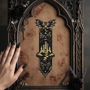 May include: A gothic-style cross-stitch design featuring a black and gold castle scene with a bat at the top. The design is framed in a dark, ornate wooden frame with decorative elements like keys and roses. The background is a tan fabric.