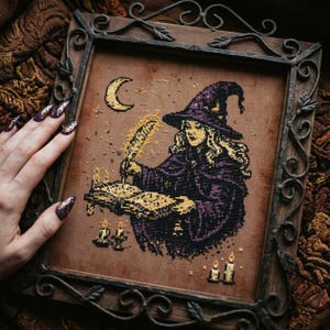 Puede incluir: Arte de punto de cruz de una bruja escribiendo en un libro, con sombrero y capa morados, una luna creciente y velas. La obra está enmarcada en un marco de madera oscuro y ornamentado con detalles metálicos. La paleta de colores incluye marrones, morados y dorados.