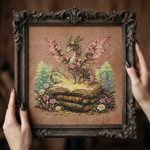 May include: Cross-stitch art of a floral dragon with pink wings and flowers, perched atop a stack of books. The artwork is framed in a dark, ornate wooden frame. The dragon is surrounded by green trees and flowers.