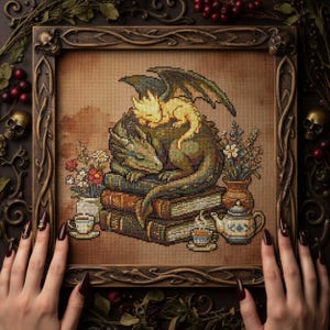 May include: Cross-stitch artwork featuring a green dragon resting on a stack of books, with a smaller yellow dragon on its back. The artwork includes flowers, teacups, and a teapot, all within a decorative wooden frame.