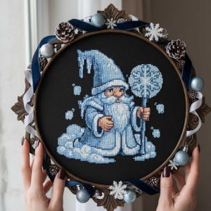 May include: A cross-stitch embroidery of a wizard in a blue robe and hat, holding a snowflake staff, set in a decorative frame. The frame is adorned with blue ribbon, ornaments, pinecones, and snowflakes, creating a winter theme.