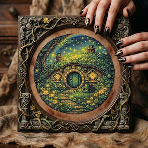 May include: A framed cross-stitch depicting a hobbit hole scene with a green door, surrounded by lush greenery and a starry sky. The frame is ornate, with a dark brown finish and decorative vines. The artwork is held by hands with black nail polish.