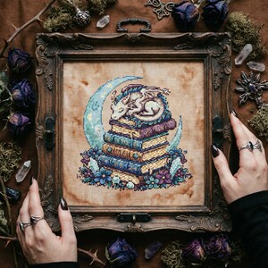 Puede incluir: Un bordado enmarcado que representa un dragón blanco durmiente sobre una pila de libros, con una luna creciente de fondo. El marco de madera está rodeado de flores secas y cristales. La paleta de colores incluye azules, morados y dorados.