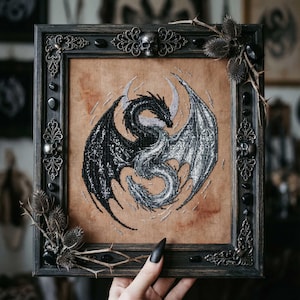May include: Framed cross-stitch art featuring two dragons, one black and one silver, intertwined against a tan background. The dark wooden frame is adorned with decorative metal accents, skulls, and black stones. Thistle and thorns are attached to the frame.