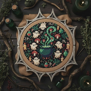 May include: A cross-stitch embroidery featuring a witchy theme. The design includes a cauldron, ghosts, mushrooms, and potion bottles in a circular frame. The color palette is dark with green, red, and white accents. The frame is a light gray color.
