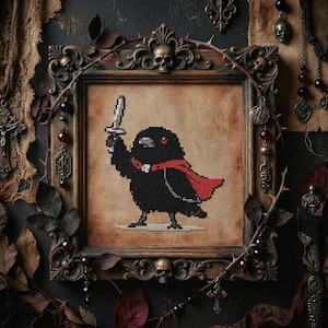 Op de afbeelding: Kruissteekkunst van een zwarte vogel met een zwaard, met een rode cape, omlijst in een decoratieve houten lijst met schedelaccenten. Het kunstwerk heeft een gotisch thema, met donkere kleuren en sierlijke details.