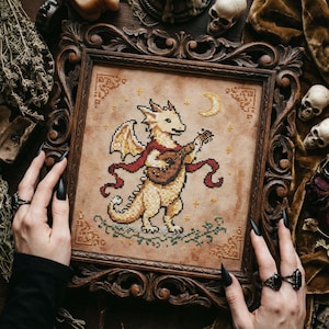 Könnte beinhalten: Kreuzstichkunst eines Drachen, der eine Laute spielt und einen roten Schal trägt, vor einem sternenklaren Nachthimmel. Das Kunstwerk ist in einem dunklen, verzierten Holzrahmen gerahmt. Der Drache ist beige und der Hintergrund ist hellbraun.