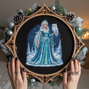 Pode incluir: Arte em ponto cruz de uma mulher em um vestido azul com uma coruja, ambientada em uma cena de inverno. A obra de arte é emoldurada em um aro de madeira decorativo, adornado com pinhas, fita prateada e enfeites. O tema é inverno e fantasia.