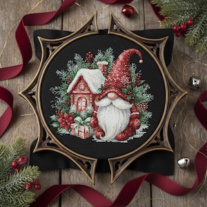Op de afbeelding: Een kruissteekborduurwerk met een kerstkabouter met een lange witte baard, een rode hoed en een rode outfit. De kabouter staat naast een met sneeuw bedekt huis en een ingepakt cadeau. De borduursel is geplaatst in een decoratieve houten lijst.