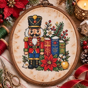 May include: A circular wooden embroidery hoop containing a cross-stitch design of a nutcracker soldier in a red and blue uniform, standing beside three books, a large red poinsettia flower, green pine branches, and a gold ornament.