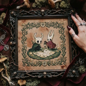 Peut inclure: Broderie représentant deux personnages du jeu vidéo Hollow Knight, encadrée dans un cadre en bois sombre et orné. L'œuvre montre les personnages sur un paysage vert et beige, entourés de vignes et de champignons verts. Le cadre a des détails de crânes.