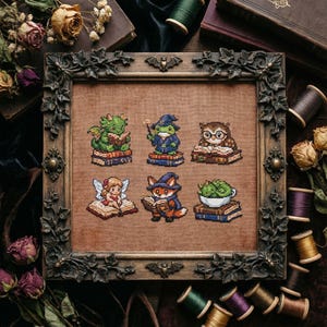 Op de afbeelding: Kruissteekkunst met zes fantasiefiguren, waaronder een draak, een kikker en een uil, die elk boeken lezen. Het kunstwerk is ingelijst in een donker, sierlijk frame met vleermuisaccenten. De achtergrond toont boeken en spoelen garen.