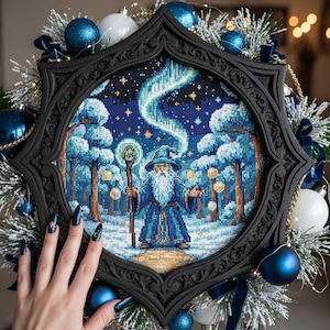 Puede incluir: Una obra de arte en punto de cruz enmarcada representa a un mago en un bosque nevado bajo la aurora boreal. El mago lleva una túnica y un sombrero azules, sosteniendo un bastón. El marco negro es adornado, rodeado de adornos azules y blancos.