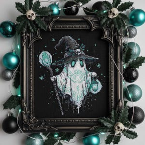 Puede incluir: Arte de punto de cruz de un fantasma con sombrero de bruja, sosteniendo un bastón y una linterna. El fantasma es blanco y verde azulado sobre un fondo negro, enmarcado en un marco oscuro y ornamentado. El marco está decorado con acebo, calaveras y adornos verde azulado y negros.