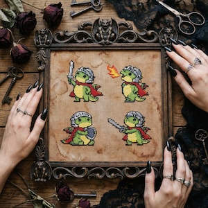 May include: Cross-stitch artwork featuring four pixelated green dragons in knight armor, each with a different action: holding a sword, breathing fire, holding a shield, and wielding a sword. The artwork is framed in a dark wooden frame with decorative carvings.