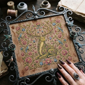 May include: A framed cross-stitch featuring a deer surrounded by flowers and birds. The embroidery uses shades of green, pink, and blue on a tan background. The frame is ornate, dark metal with decorative birds and roses.