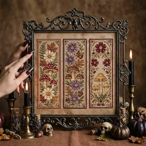 Puede incluir: Un bordado de punto de cruz rectangular que presenta tres paneles verticales con motivos botánicos y de setas, enmarcado en un marco de metal oscuro ornamentado con un detalle de murciélago en la parte superior. Los paneles muestran setas y flores.