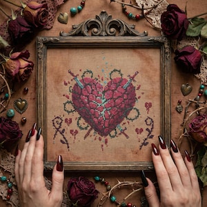May include: A framed cross-stitch featuring a heart pierced by two swords, surrounded by thorny vines and small hearts. The heart is a deep red color, with blue accents. The frame is a dark brown color.