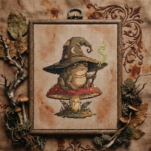 May include: Cross-stitch art of a frog wizard wearing a pointed hat with a crescent moon and stars, sitting on a red mushroom with white spots. The frog holds a wand with green smoke. The artwork is in a wooden frame.