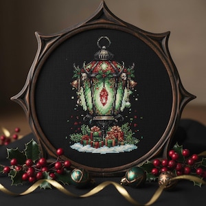 May include: A framed cross-stitch depicting a festive lantern. The lantern is green and red, decorated with holly and skulls. Presents and snow are at the base. The dark brown frame surrounds the black background.