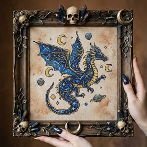 May include: A framed cross-stitch depicting a blue and gold dragon with celestial motifs. The artwork is set against a tan backdrop and framed in a dark, ornate frame with skulls, crystals, and crescent moons.
