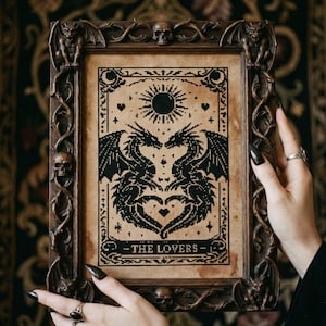 May include: A framed cross-stitch art piece featuring two black dragons facing each other, forming a heart shape, with a sun above. The artwork is labeled "THE LOVERS" and is set in a dark brown, ornate frame with skulls and gargoyles.