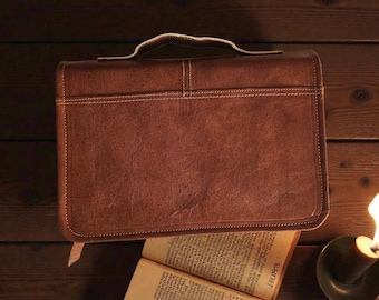 Puede incluir: Una funda de Biblia de cuero marrón con asa y costuras blancas. La funda es rectangular y está sobre una superficie de madera junto a un libro abierto y una vela encendida.