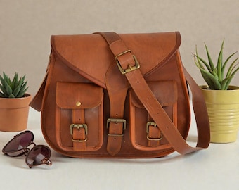 Könnte beinhalten: Eine braune Ledertasche mit zwei Vordertaschen, die jeweils mit einer Schnalle verschlossen sind. Die Tasche hat einen Klappverschluss und einen langen, verstellbaren Riemen. Sonnenbrillen und Topfpflanzen sind im Hintergrund.
