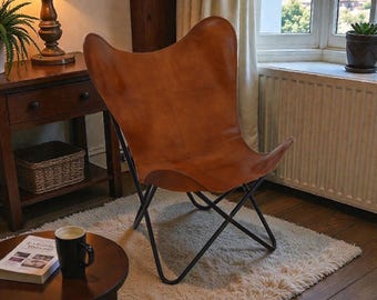 May include: A brown leather butterfly chair with a black metal frame. The chair is set on a fluffy white rug. A small wooden side table with a lamp and a book and mug are in the foreground. The room has a warm, inviting atmosphere.