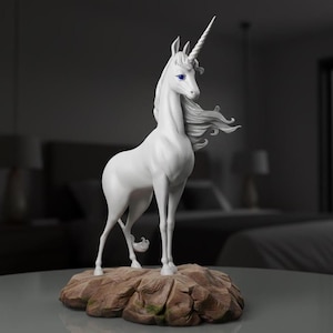 May include: A white unicorn ornament with a spiralled horn and flowing mane, standing on a brown, rocky base. The unicorn has blue eyes and is posed gracefully. The background is blurred, suggesting a room setting.