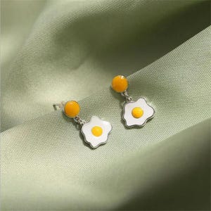 May include: Silver-coloured earrings with a fried egg design. Each earring has a white egg shape with a yellow yolk and a yellow circle at the top. The earrings are set against a light green background.