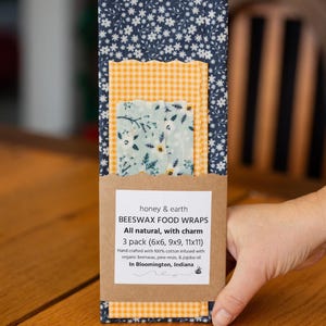 May include: A three-pack of beeswax food wraps featuring a dark blue floral print, a yellow and white gingham check, and a light blue floral pattern. The packaging includes the text honey & earth BEESWAX FOOD WRAPS 3 pack (6x6, 9x9, 11x11).