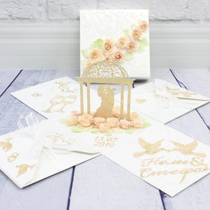 May include: A white pop-up greeting card featuring a gold gazebo with a couple silhouette inside, surrounded by peach-colored paper roses. The card displays gold foil illustrations of champagne flutes, doves, and the text 13.07.2019 and names.