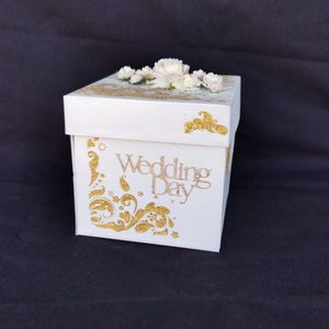 May include: A white square gift box with gold detailing and the words "Wedding Day" in gold script. The box is decorated with gold floral designs and topped with white and pink flowers.