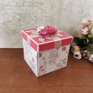May include: A square gift box with a white base featuring a delicate pink and grey floral pattern. The lid is solid pink with a light pink ribbon accent and a cluster of artificial pink and white flowers attached to the top centre of the lid.